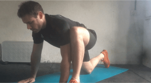Fitness instructor demonstrates the first stage of the hip opener stretch by assuming a lunch position with one knee touch the floor behind and one foot extended forward. Hands are palm facing down on the floor next to the front foot. 