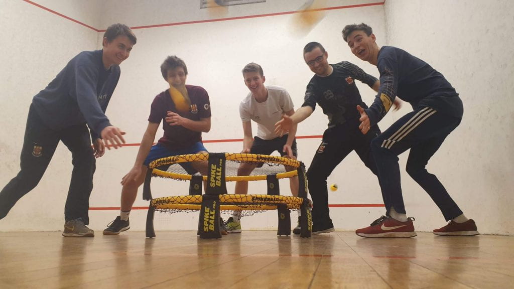 Five male students a stood in a squash court, gathered in a semi-circle around a roundnet. The roundnet is yellow and resembles a small trampoline. The group look like they are having fun.