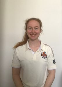 Nadia, a woman in a short sleeved cricket polo top, stands smiling against a white background. She is the treasurer of UBWCC Cricket Club.